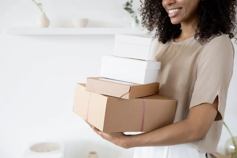 Woman with curly hair carrying stack of packaging boxes in a bright room