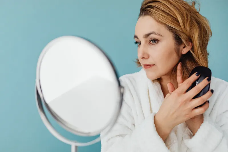 A woman brushes her hair in front of a mirror with a pastel blue background